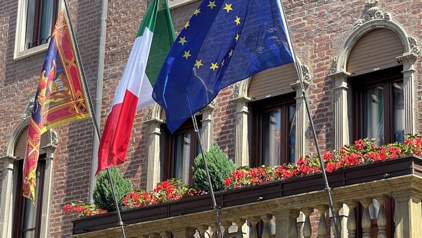 Rakennuksessa on useita lippuja, muun muassa Italian ja Euroopan unionin liput, ja sen parvekkeella on punaisia kukkia.