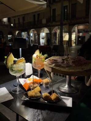 A table with a variety of food items and drinks, including a glass with a lime wedge and mint, a plate with bread and meat, and a dessert on a stand. - Hotel Serafino  (Padova)