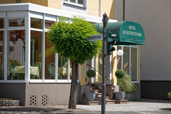 A hotel entrance with a green awning labeled "HOTEL KURFÜRSTENHOF" and a street sign for "Meckenheimer-Allee".