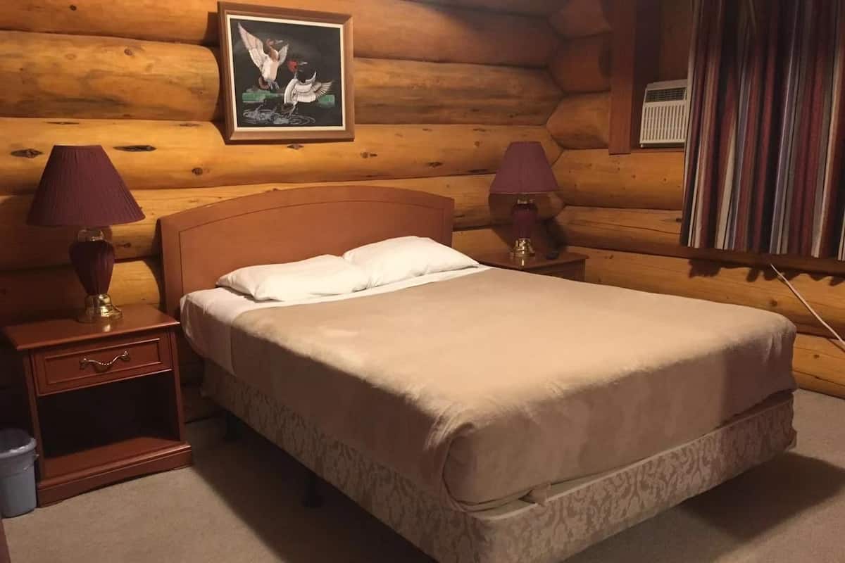 A cozy log cabin bedroom with a wooden bed, two bedside lamps, a framed picture on the wall, and a window with curtains.