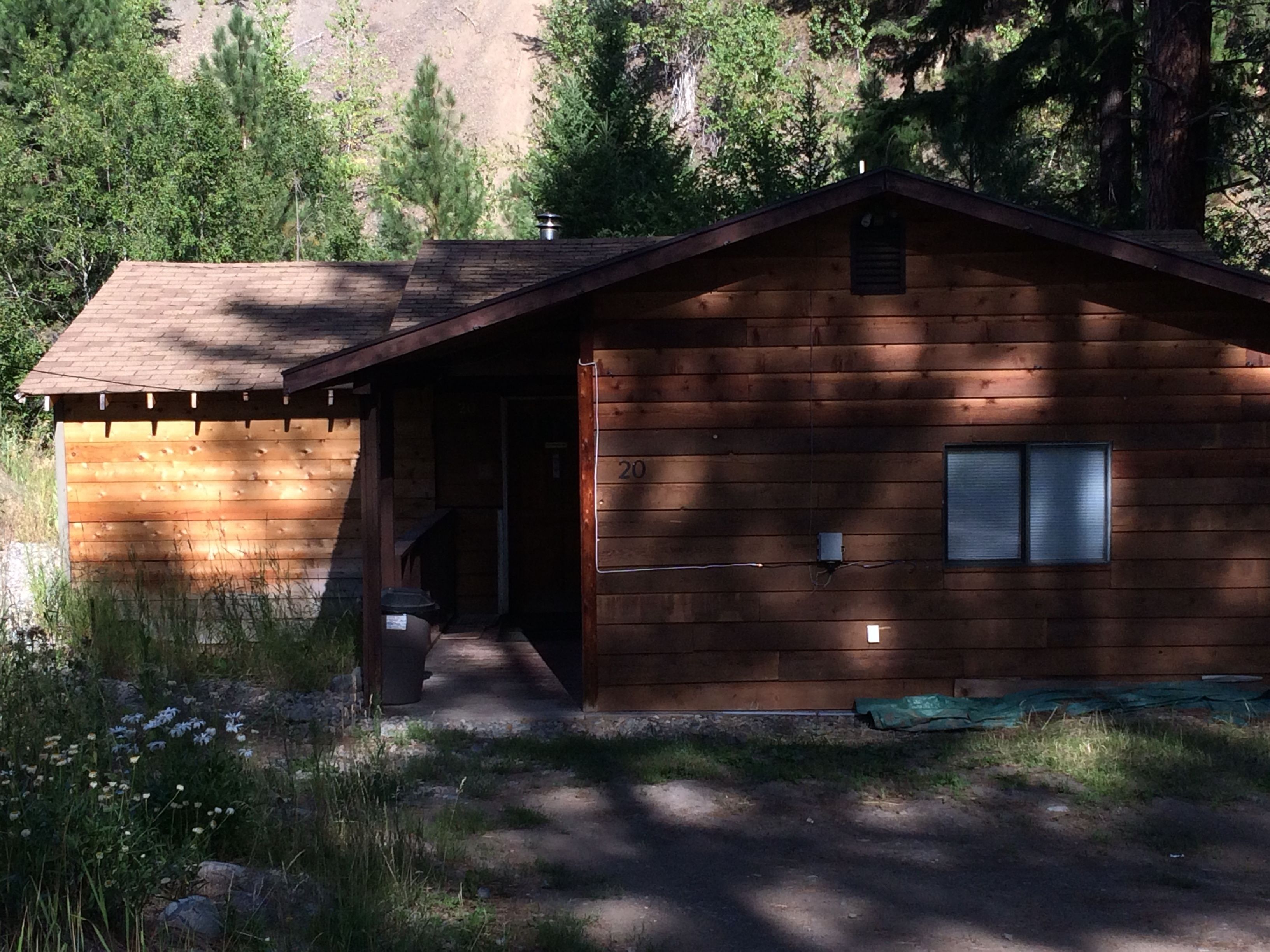 Une cabane en bois avec une seule porte et une petite fenêtre, entourée d’arbres et bordée d’un chemin de terre.