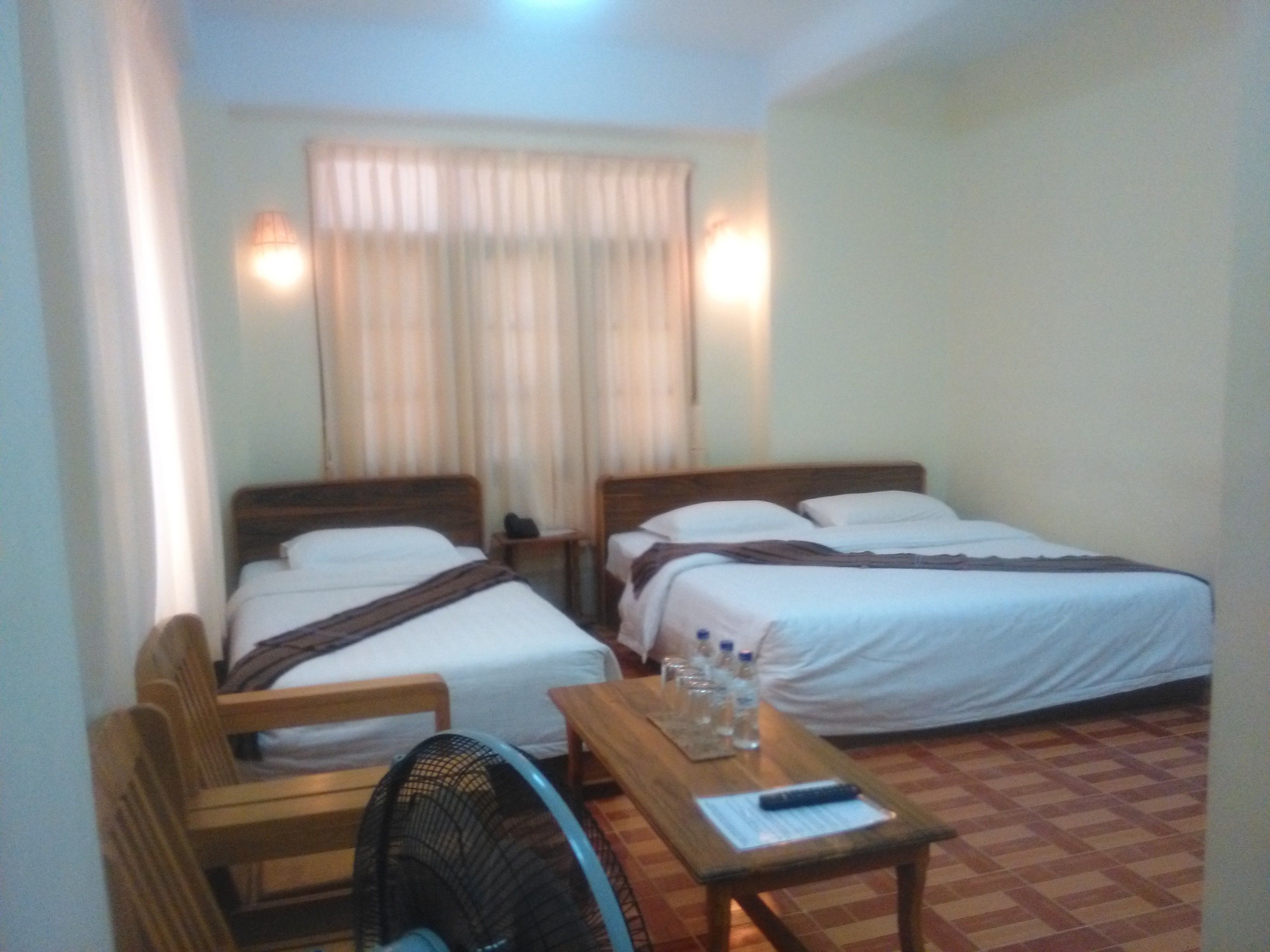 A hotel room with two single beds, a wooden chair, a table with a fan, and a window with curtains.
