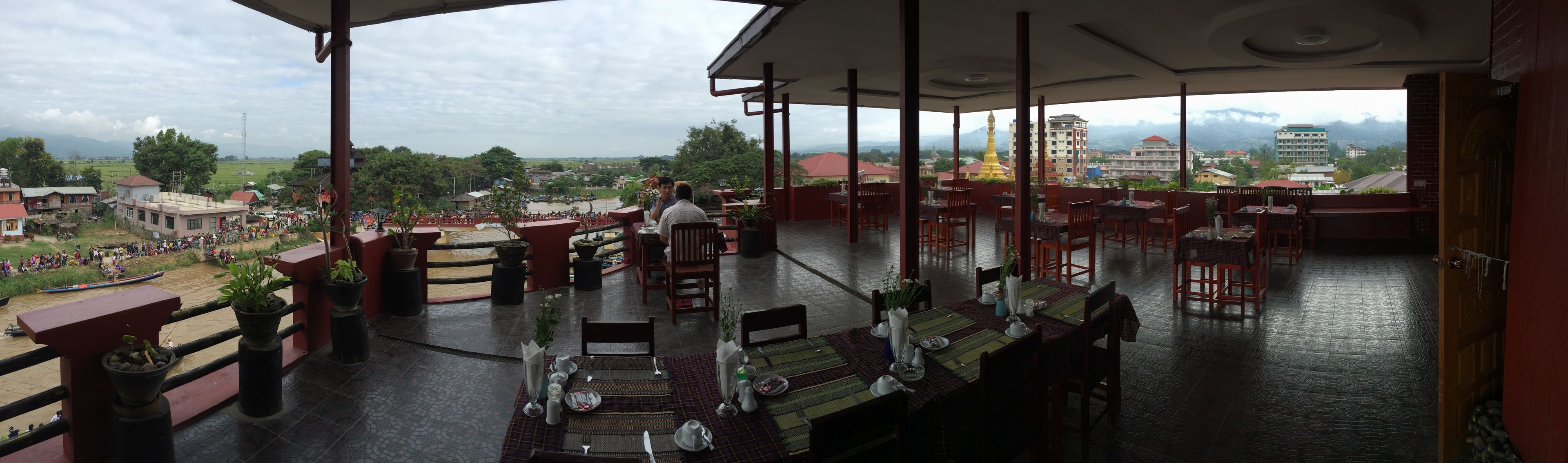 A restaurant with tables set for dining, a view of a river with boats, and a distant view of a golden temple and buildings.