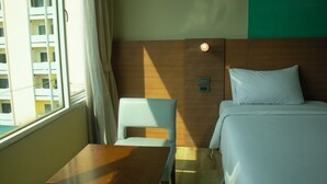 Une chambre d’hôtel avec un lit, une petite table, une chaise, une fenêtre avec des rideaux et un luminaire fixé au mur.