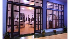 Entrada de hotel con puerta de cristal, zona de comedor visible en el interior y plantas en macetas en el exterior.