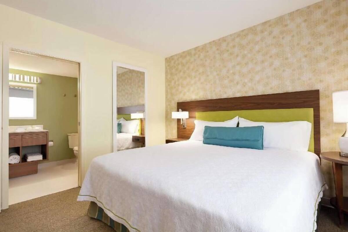 Quarto de hotel com uma cama grande, banheiro com vaso sanitário e pia, e um espelho.