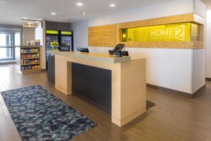 A hotel reception desk with a 'HOME 2 Suites by Hilton' sign, a self-service kiosk, and a snack bar with a variety of snacks. - Home2 Suites by Hilton Gillette (Gillette)