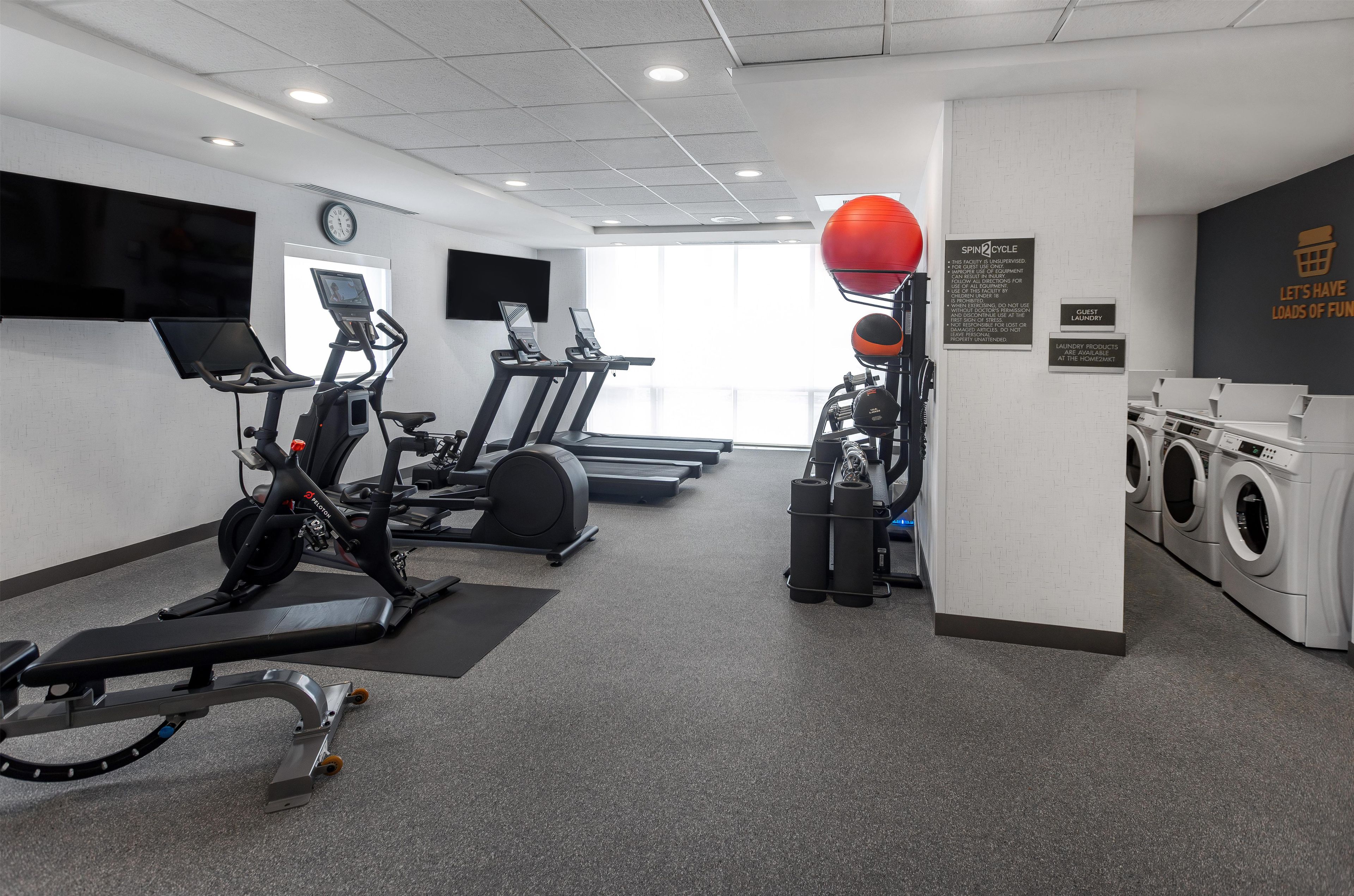 Une salle de sport équipée de tapis de course, de vélos d’appartement, d’un appareil d’aviron, d’un ballon d’exercice rouge et d’un espace buanderie.