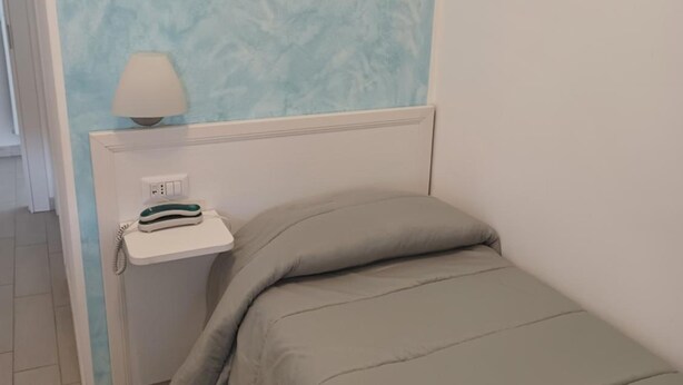 A small, neatly made room with a bed, a bedside table with a lamp, and a wall with a textured pattern.