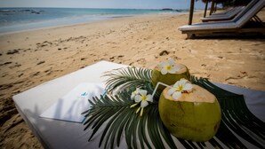 Escena de playa con una mesa dispuesta para dos, mostrando un mantel blanco, dos cocos adornados con flores y una hoja de palma.