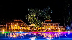 Vista nocturna de un hotel con piscina, mobiliario exterior y un gran árbol.