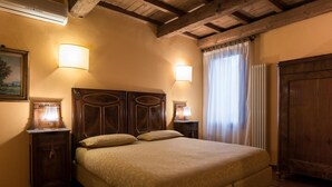 A bedroom with a large bed, wooden headboard, two bedside tables with lamps, a radiator, and a window with curtains. - Piazza Nova Guest House (Ferrara)