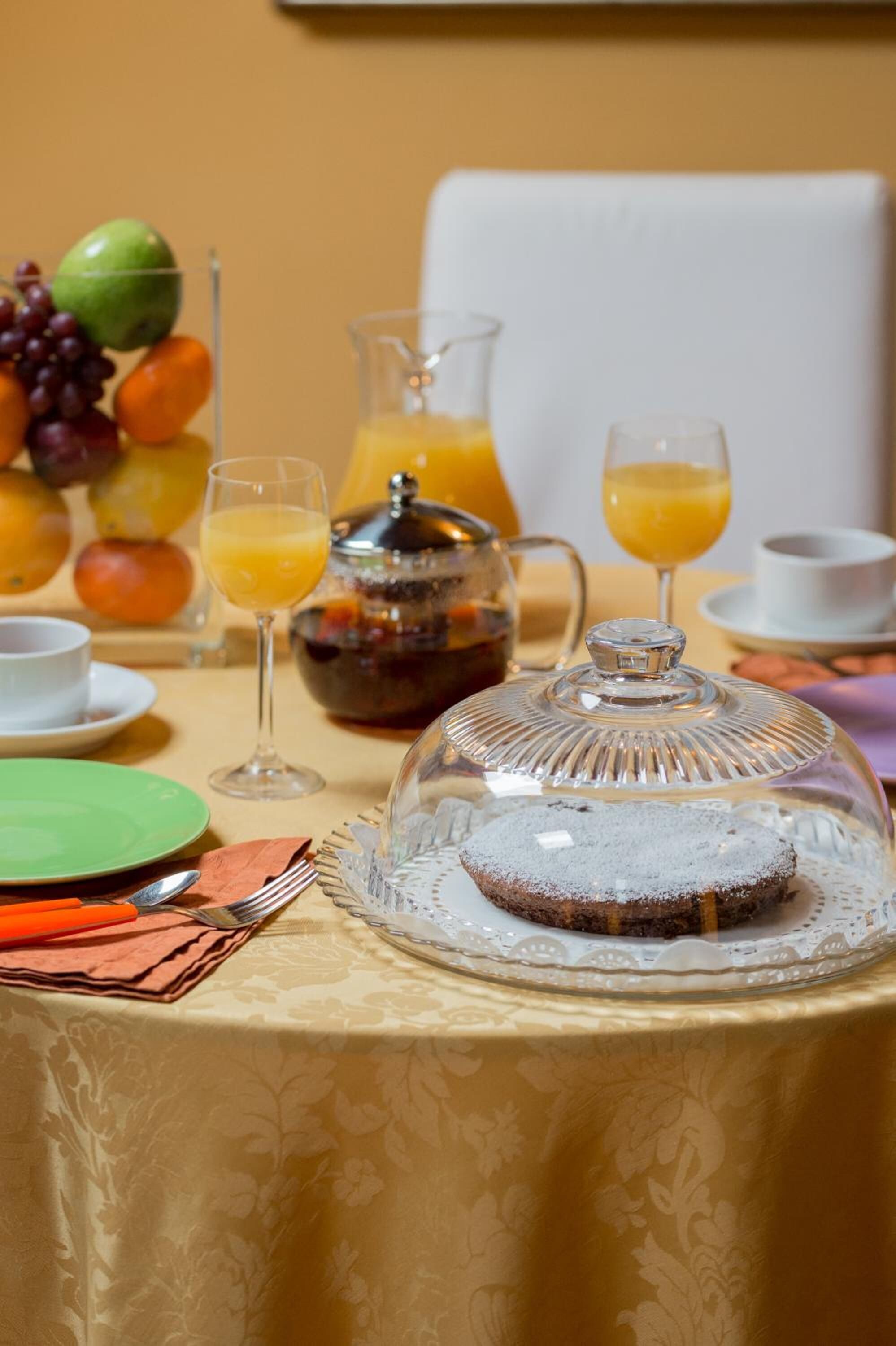A breakfast setting with a fruit arrangement, a coffee pot, orange juice glasses, and a covered dessert on a table.