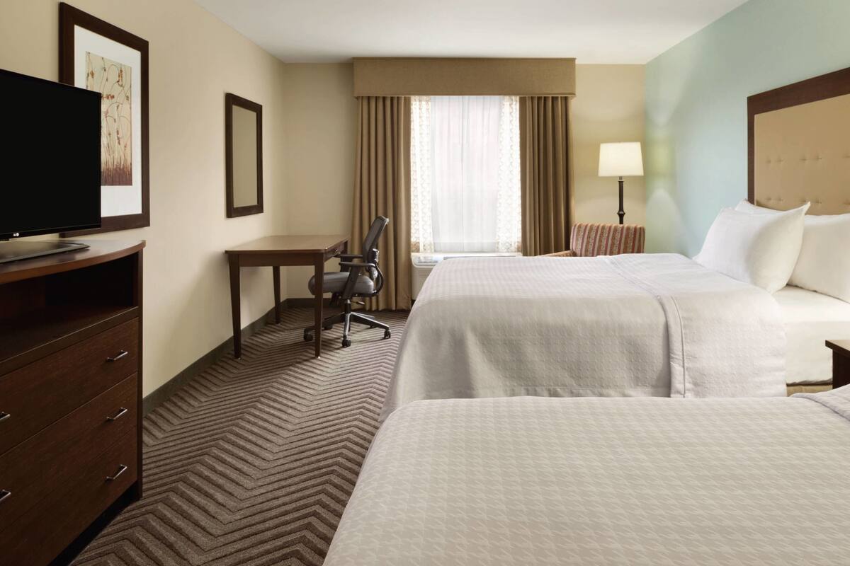 A hotel room with two beds, a desk, a chair, a TV, and a window with curtains.
