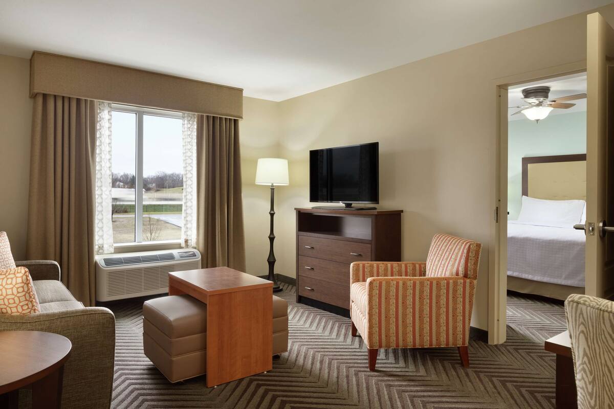A hotel room with a large window, a flat-screen TV, a desk, a chair, a bed, and a ceiling fan.