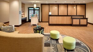 A lobby with a reception desk, a sofa, two ottomans, and a small table with decorative items. - Homewood Suites by Hilton Kalamazoo-Portage (Portage)