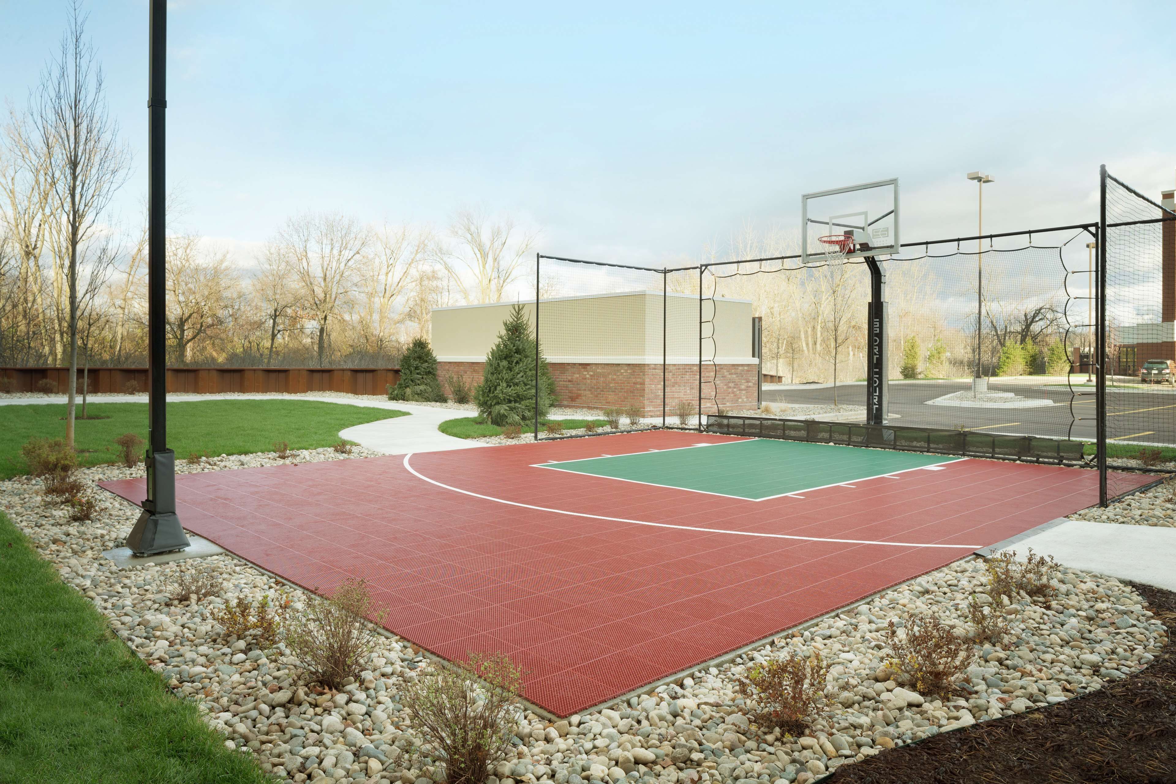 An outdoor basketball court with a red surface, surrounded by a gravel border and small plants.