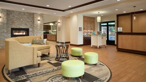 A lobby with a sofa, ottomans, a fireplace, a bar area, and a refrigerator stocked with beverages. - Homewood Suites by Hilton Kalamazoo-Portage (Portage)