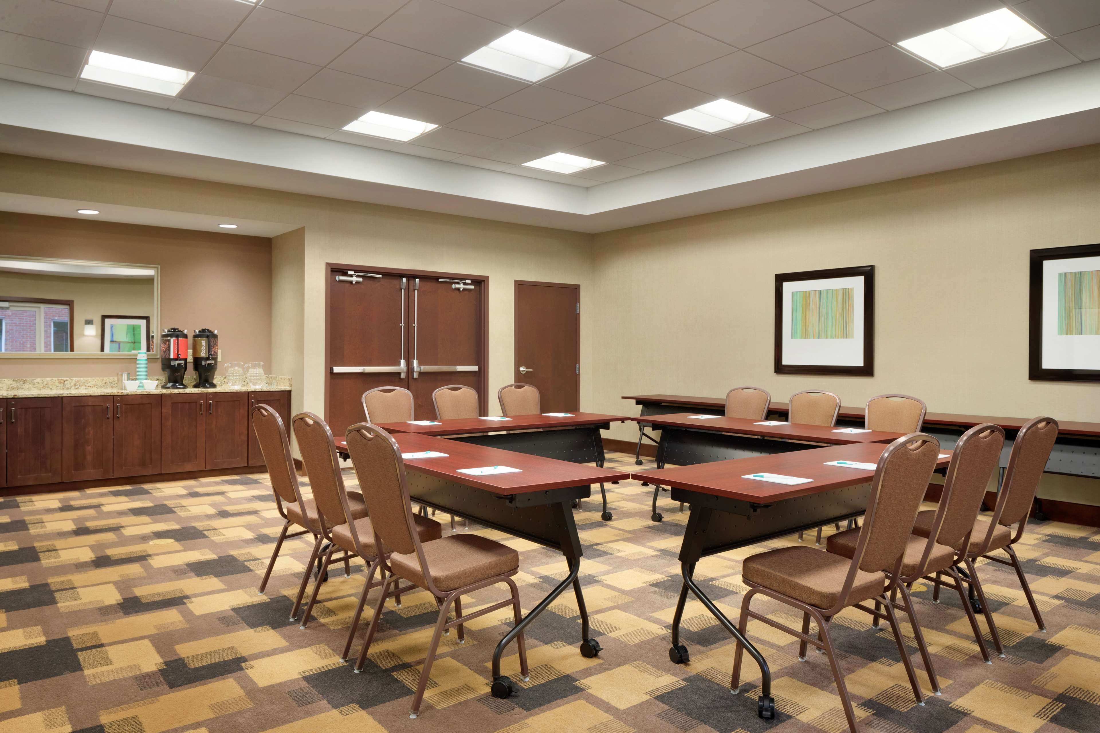 A conference room with a checkered floor, brown chairs, and tables. There are framed pictures on the walls and a kitchenette area in the background.