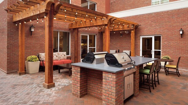 An outdoor patio with a brick barbecue area, wooden pergola, and seating. - Homewood Suites by Hilton Kalamazoo-Portage (Portage)