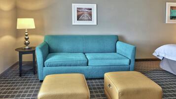 A hotel room with a teal sofa, a small table with a lamp, a framed picture on the wall, and a bed with a white cover.