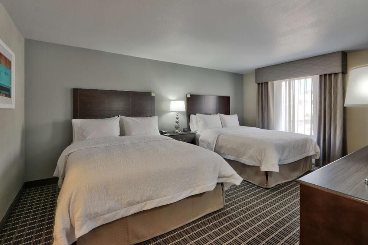 A hotel room with two beds, a desk, a lamp, and a window with curtains.
