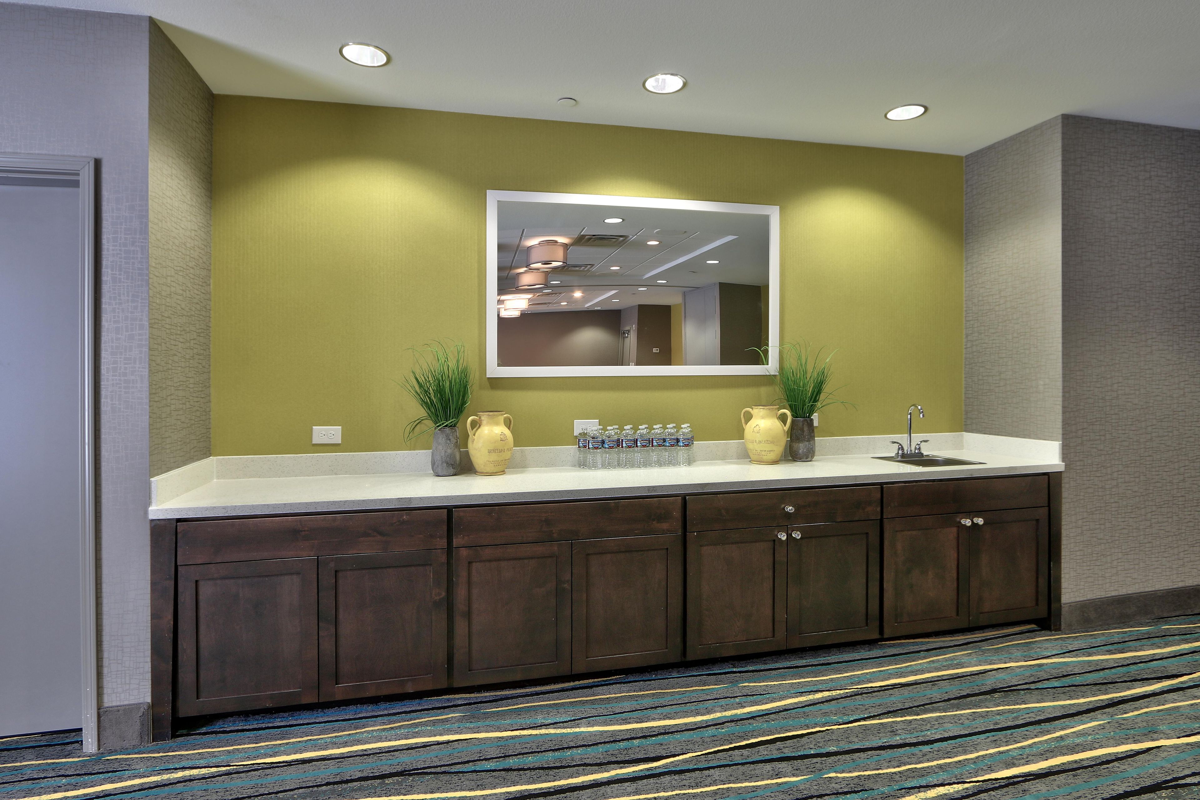 A hotel bathroom with a large mirror, dark wooden cabinets, a sink, and a patterned carpet.