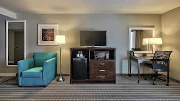 A hotel room with a teal armchair, a flat-screen TV, a minibar, a desk with a chair, and a large mirror.