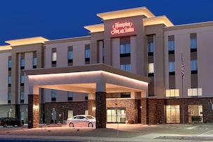 Un hotel Hampton Inn & Suites con entrada techada, varios pisos y una bandera estadounidense.