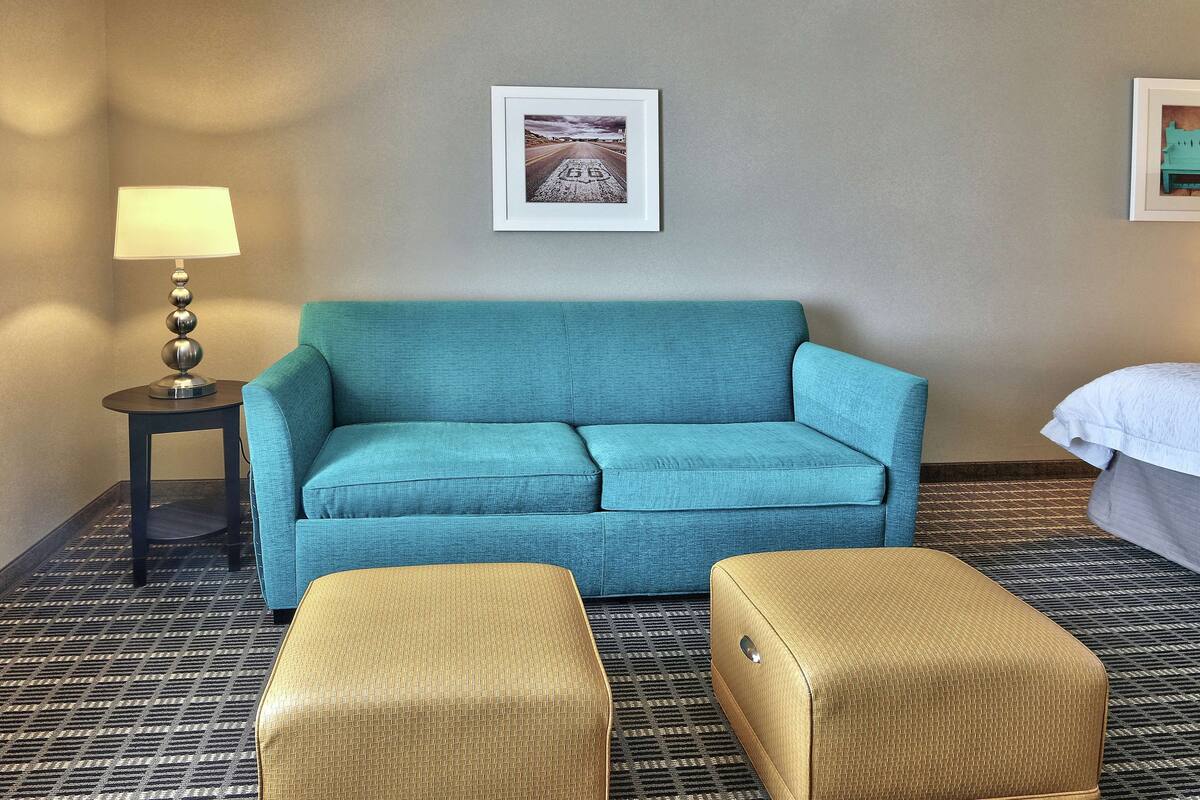Camera d'albergo con un divano color petrolio, due poggiapiedi dorati, un tavolino con una lampada e un quadro appeso alla parete.