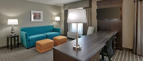 A hotel room with a sofa, ottoman, desk, lamp, and a flat-screen TV.