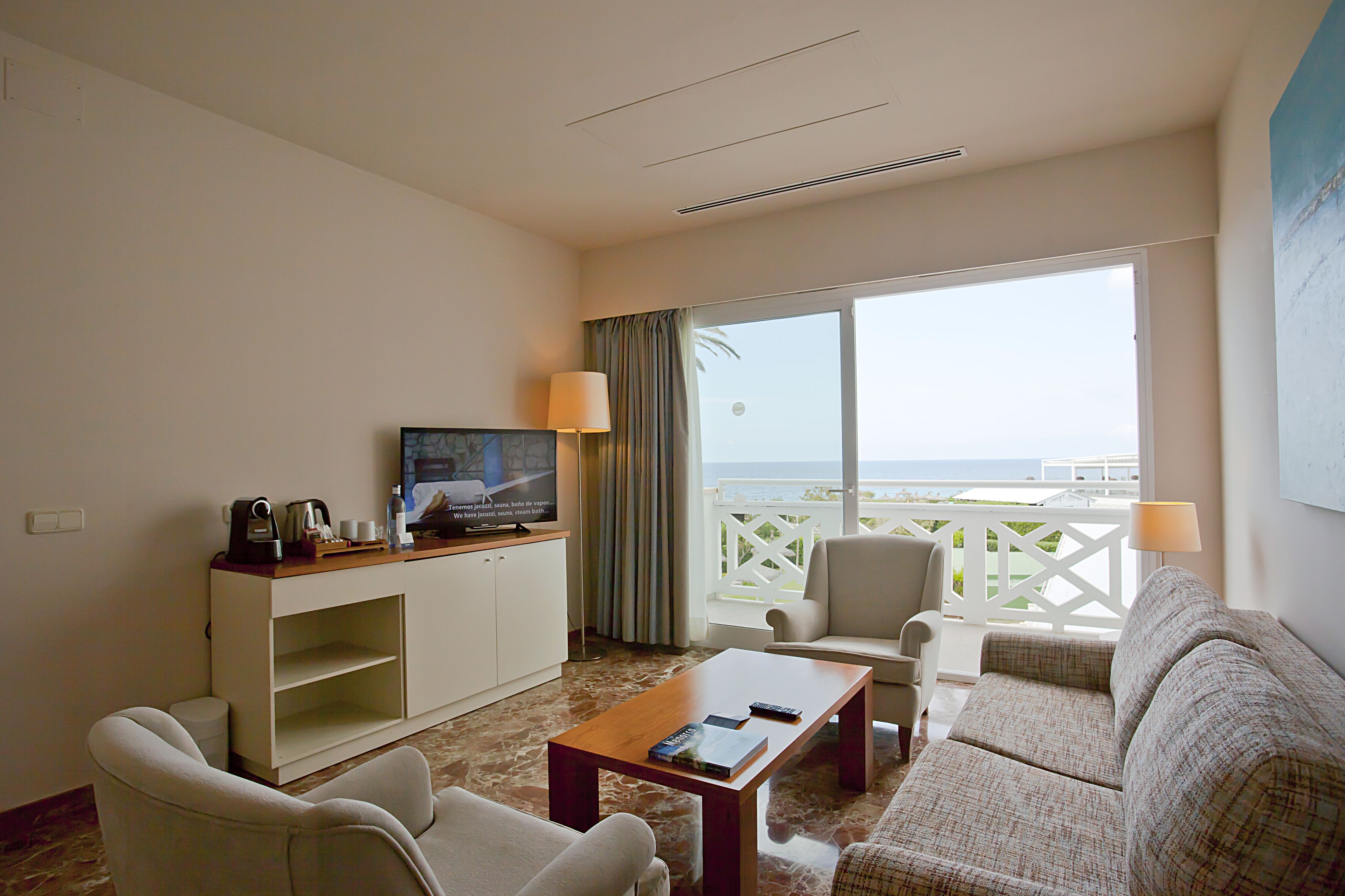 A hotel room with a balcony, a television, a sofa, a coffee table, and a desk with a lamp.