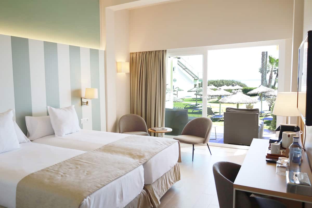 A hotel room with two beds, a desk, a chair, and a view of the beach and palm trees.