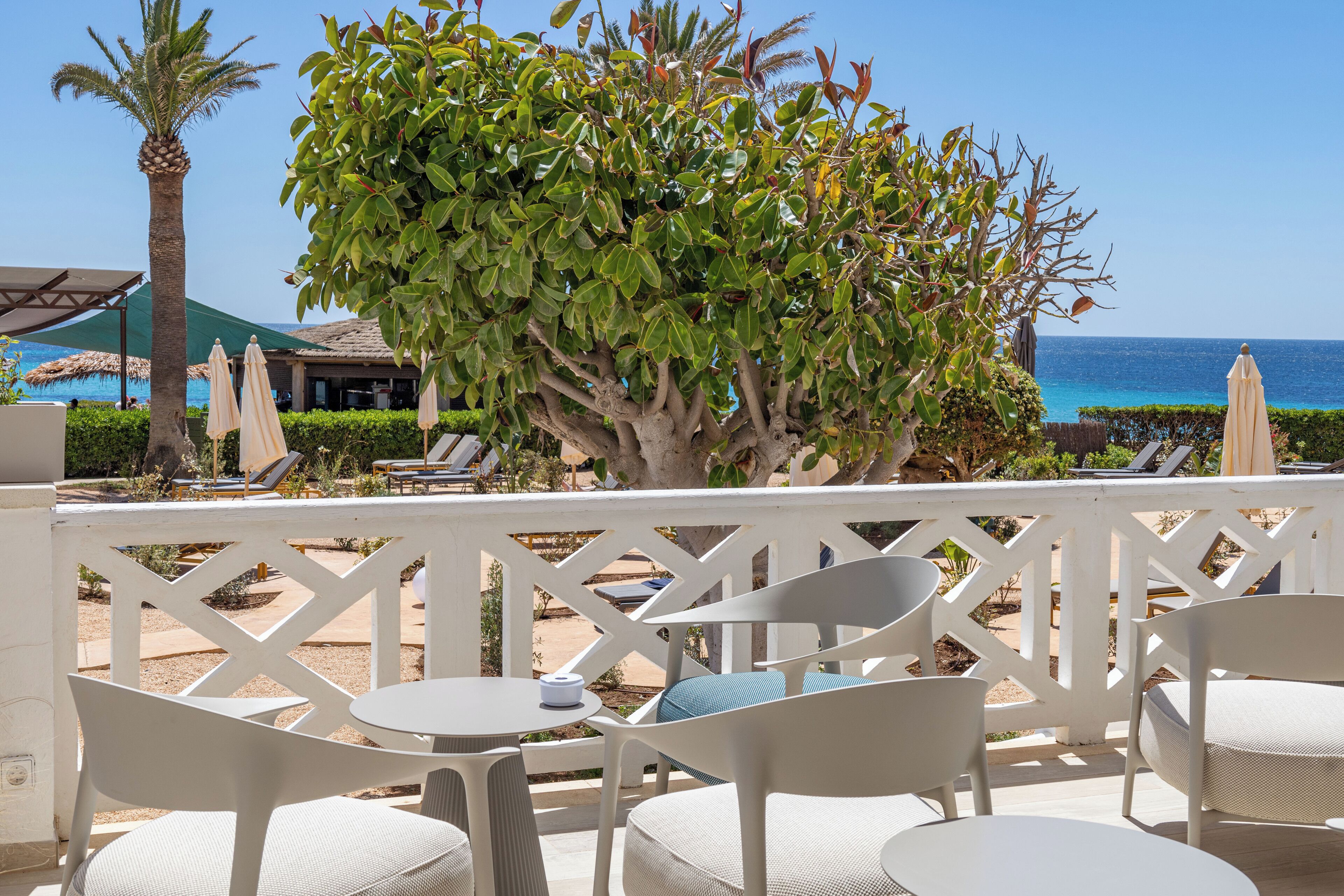A terrace with white chairs and tables, overlooking a beach with umbrellas and lounge chairs.