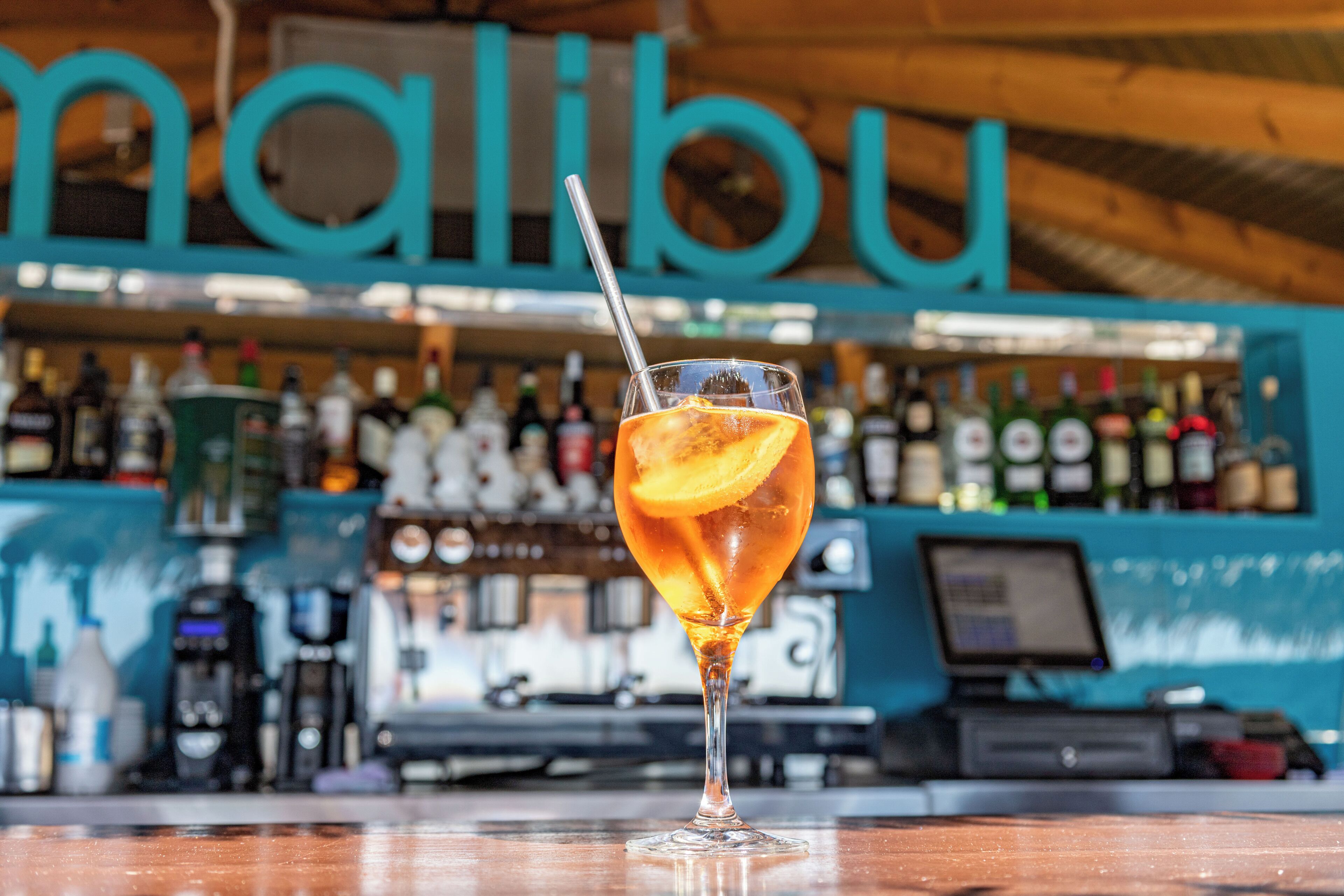 A bar with a glass of amber-colored drink with an orange slice and a straw, a cash register, and shelves stocked with bottles.