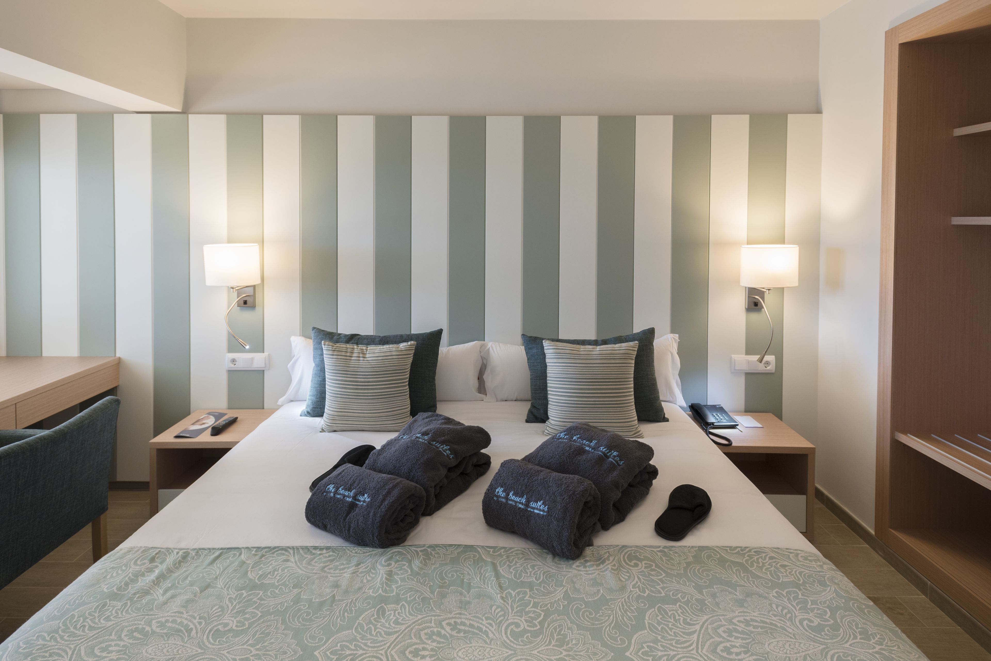 A hotel room with a bed, two bedside tables, a desk, and a chair. There are towels and slippers on the bed, and a phone on the desk.