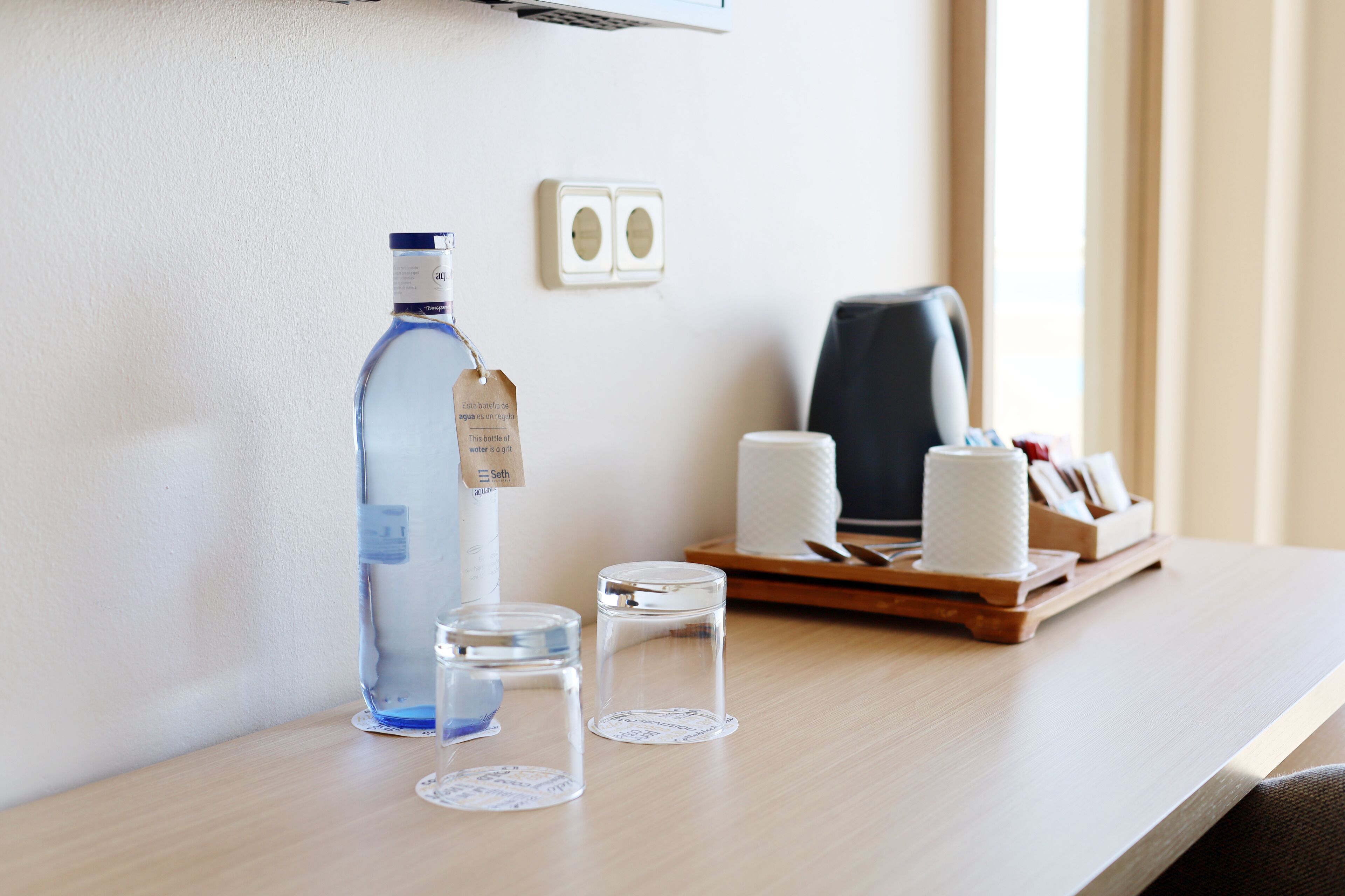 A hotel room with a wooden desk, a bottle of water, two glass containers, a tissue roll, and a tray with a tissue box and a pen.