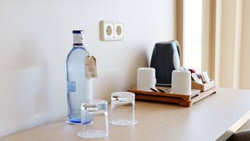 A hotel room with a wooden desk, a bottle of water, two glass containers, a tissue roll, and a tray with a tissue box and a pen.