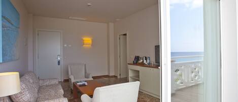 A hotel room with a balcony, a sofa, a chair, a table, and a desk with a lamp.