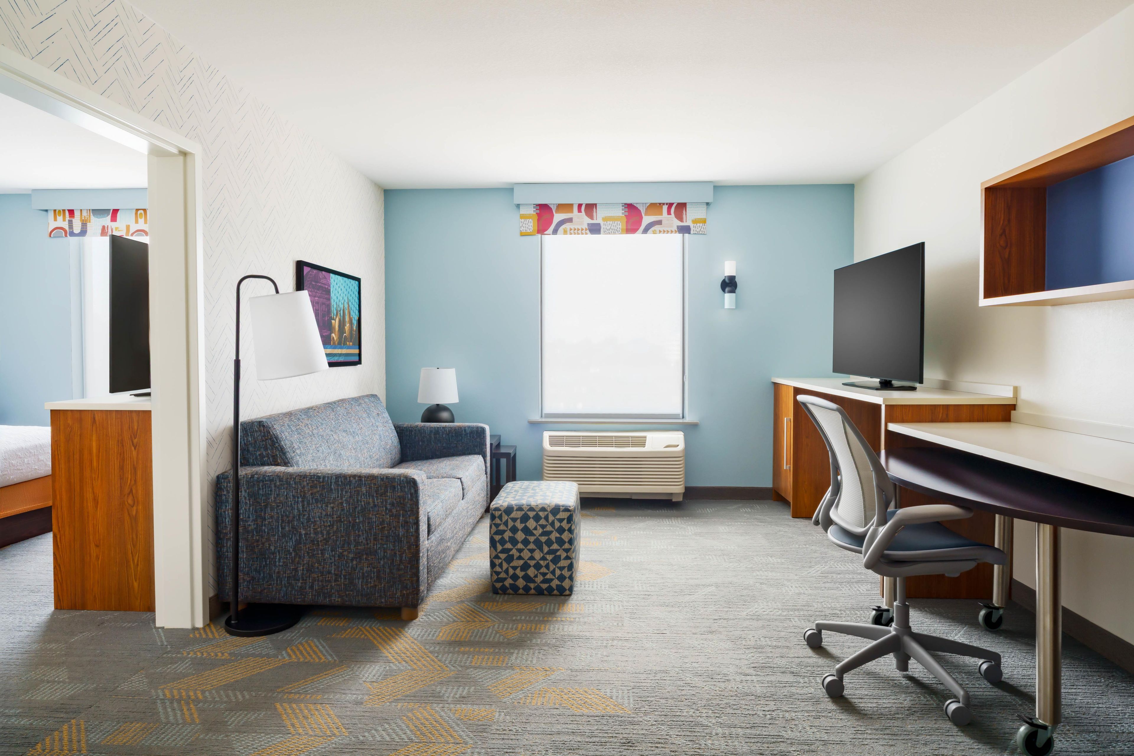 A hotel room with a bed, a sofa, a desk with a computer, a TV, and a window with curtains.