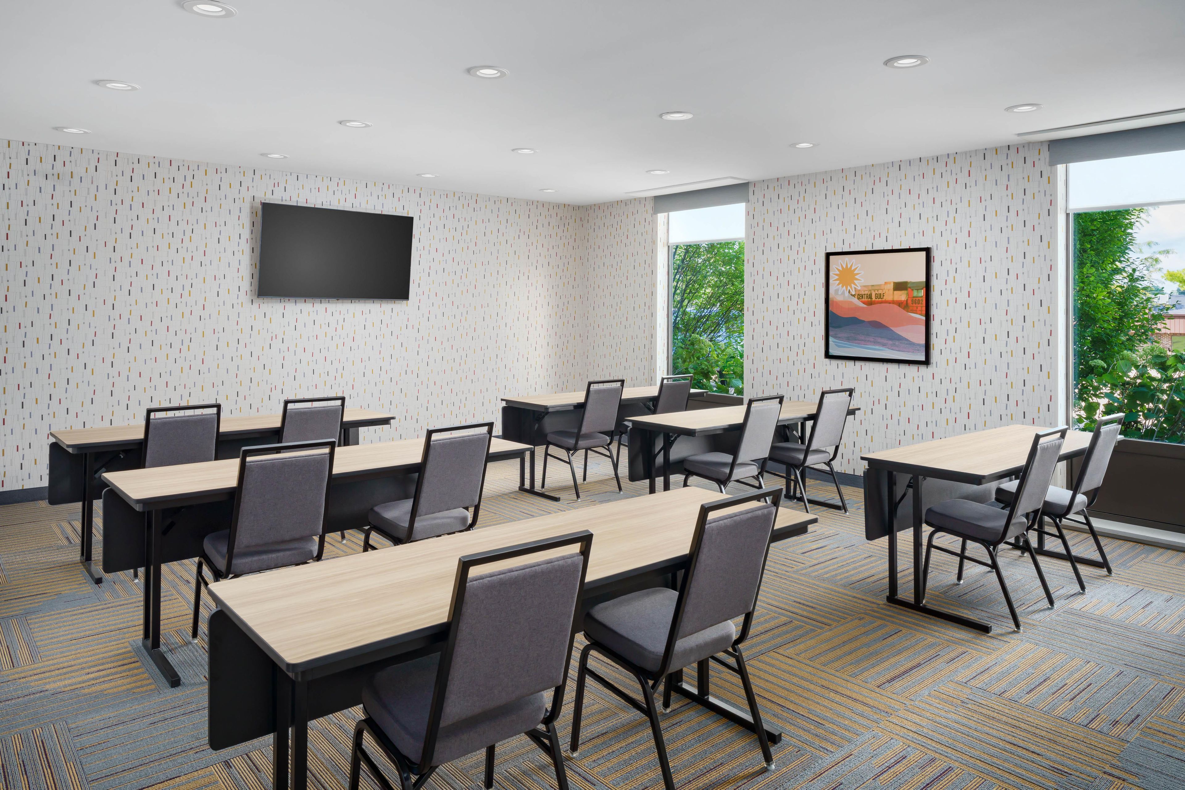 A conference room with multiple tables and chairs, a flat-screen TV mounted on the wall, and a large window providing a view of greenery outside.