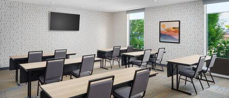 A conference room with multiple tables and chairs, a flat-screen TV mounted on the wall, and a large window providing a view of greenery outside.