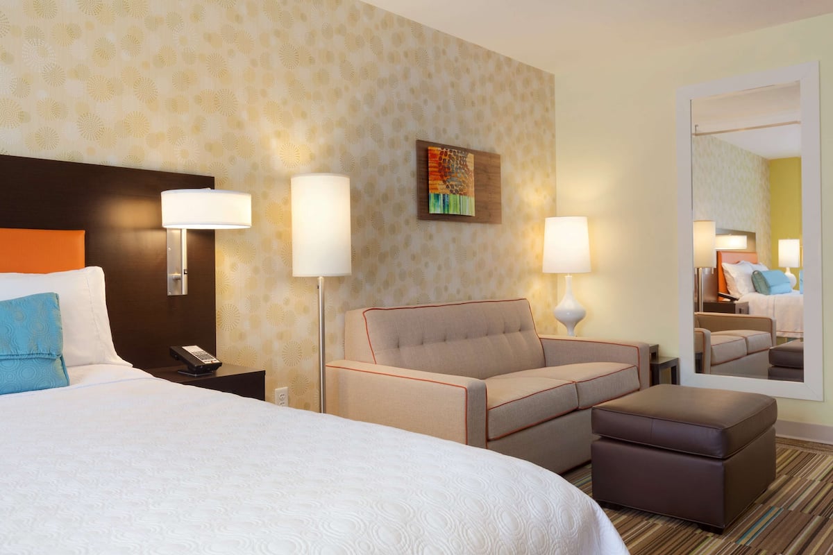 A hotel room with a large bed, a sofa, an ottoman, a nightstand with a phone, a lamp, and a painting on the wall.