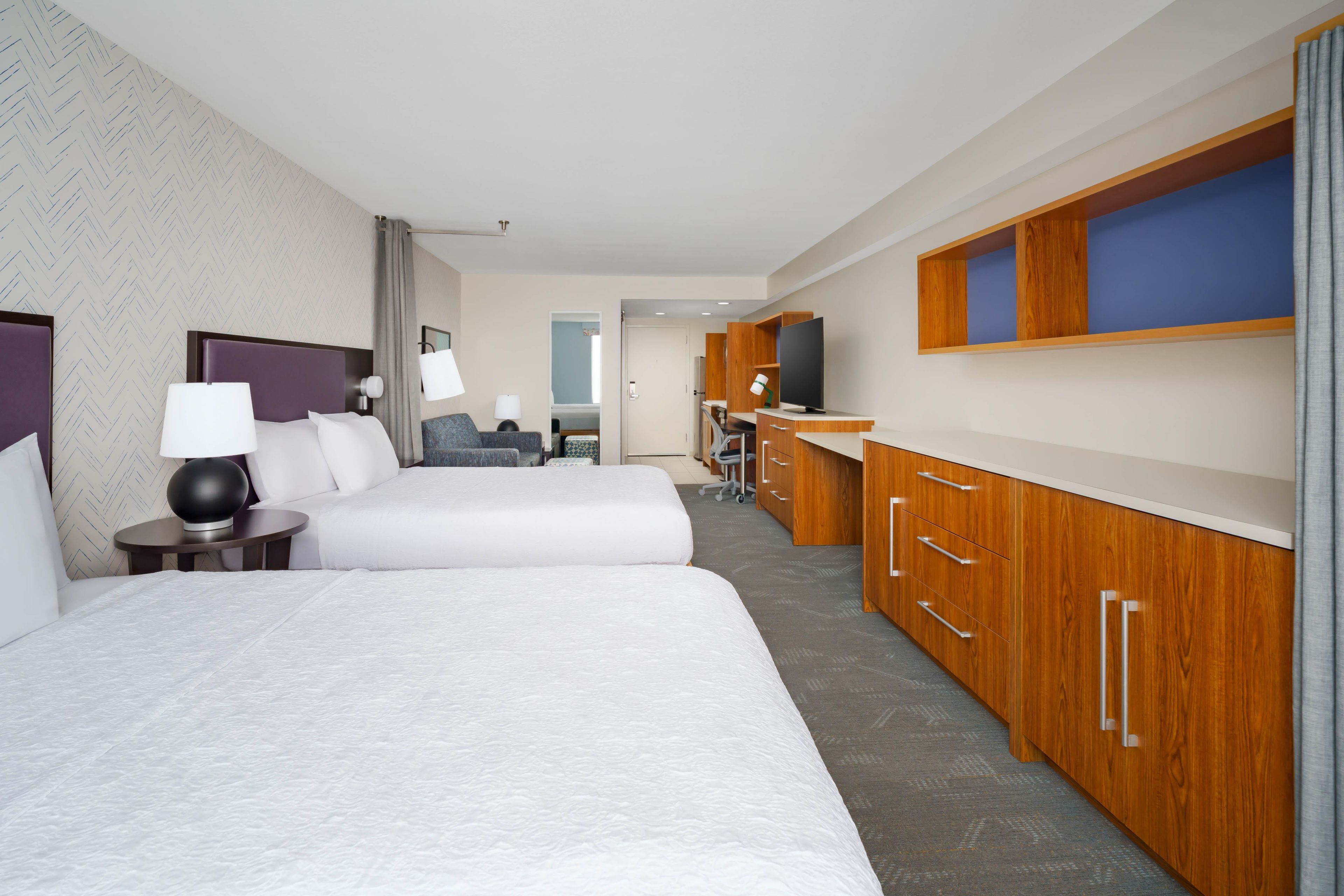 A hotel room with two beds, a desk, a TV, and a minibar.