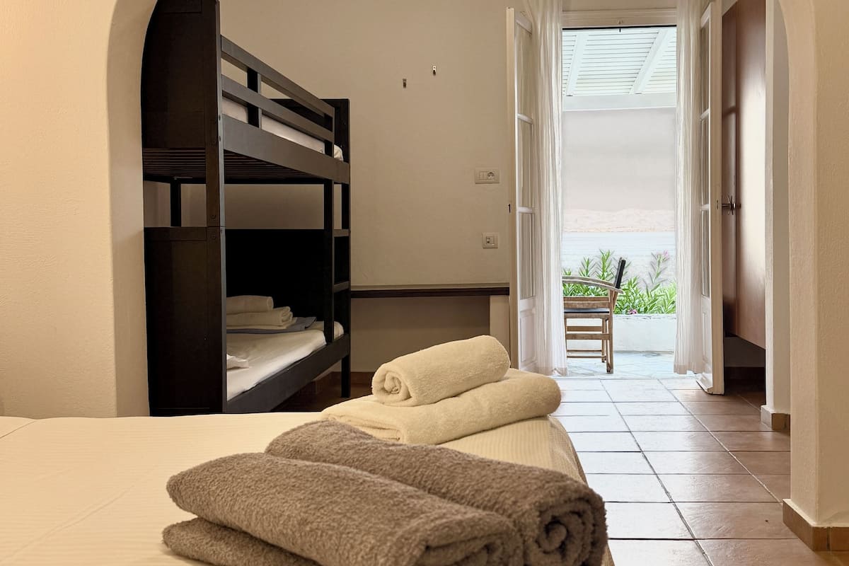 A room with a bunk bed, a bed with rolled towels, and an arched doorway leading to a balcony with plants.
