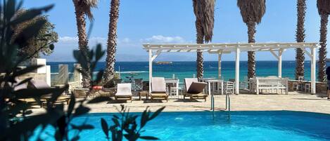Zona de piscina con sillas de descanso y una pérgola, rodeada de palmeras con vista al océano.