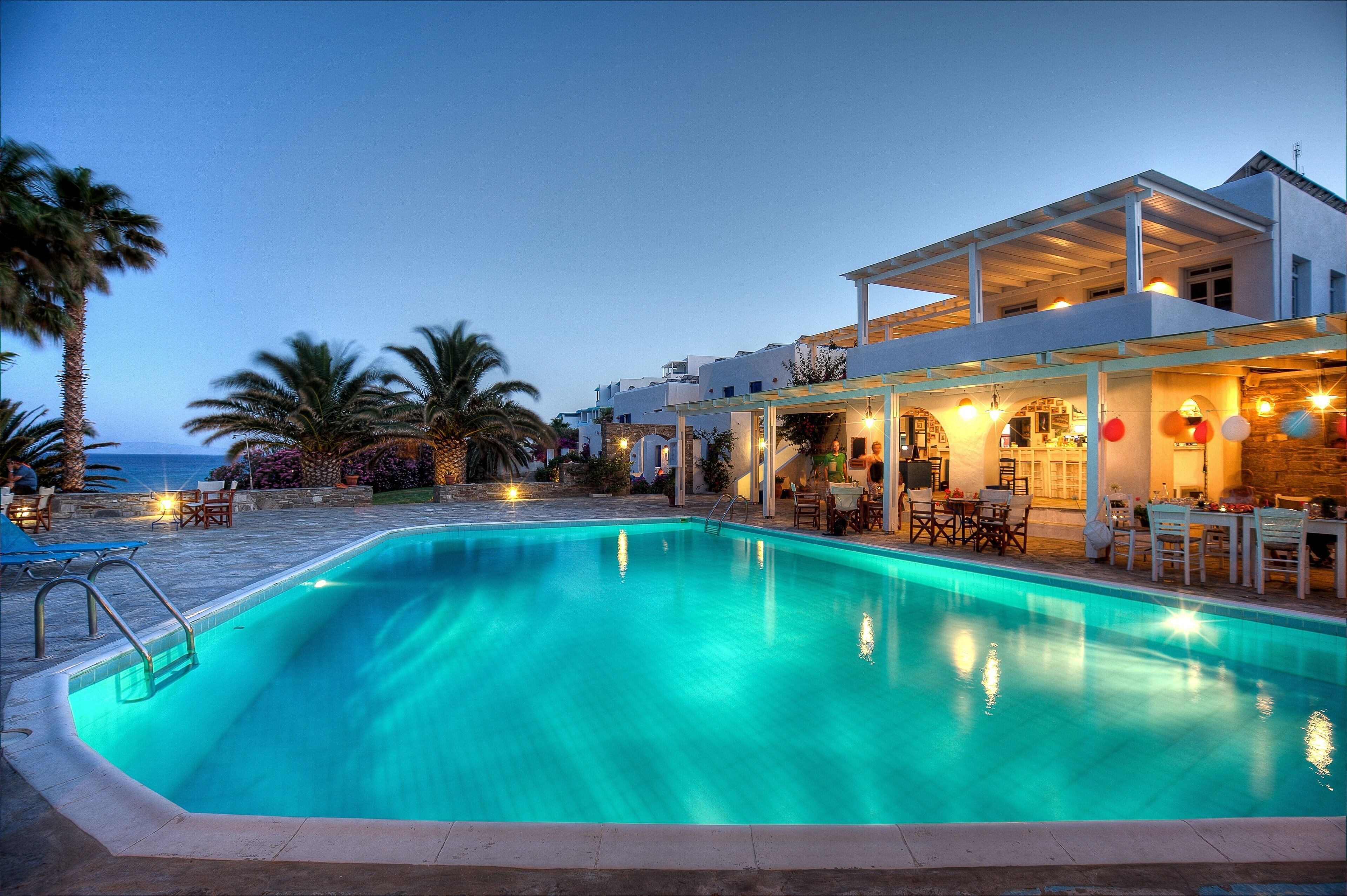 A swimming pool with a clear blue water, surrounded by lounge chairs and tables, and a building with a covered patio area.
