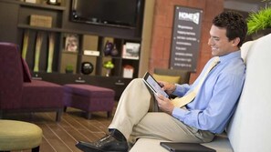 A man sitting on a couch in a modern lobby with a flat-screen TV mounted on the wall, a reception desk, and a signboard. - Home2 Suites by Hilton Atlanta South/McDonough (McDonough)