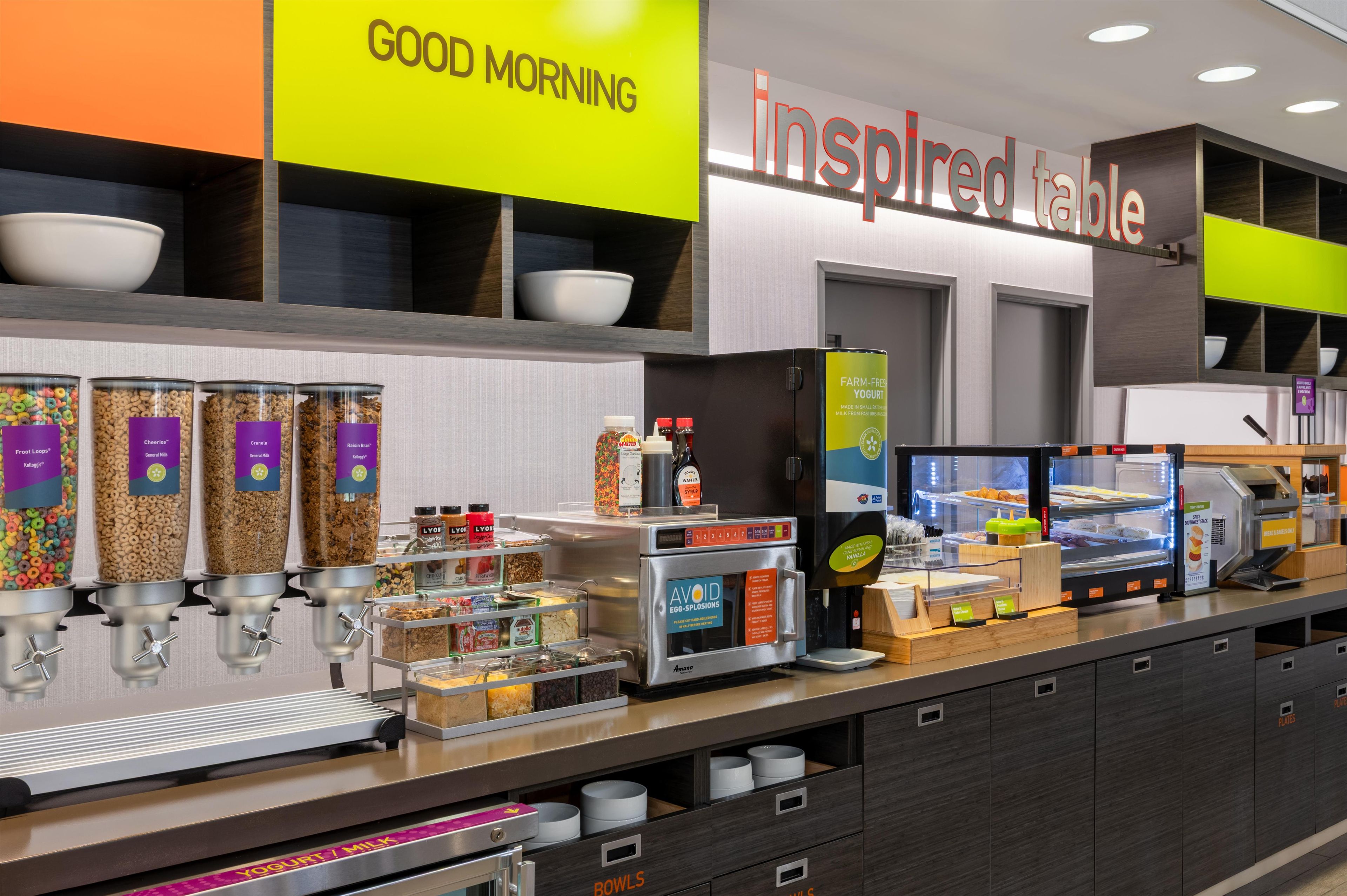 Una barra de desayuno con varios dispensadores de comida, una máquina de café y un mostrador refrigerado.