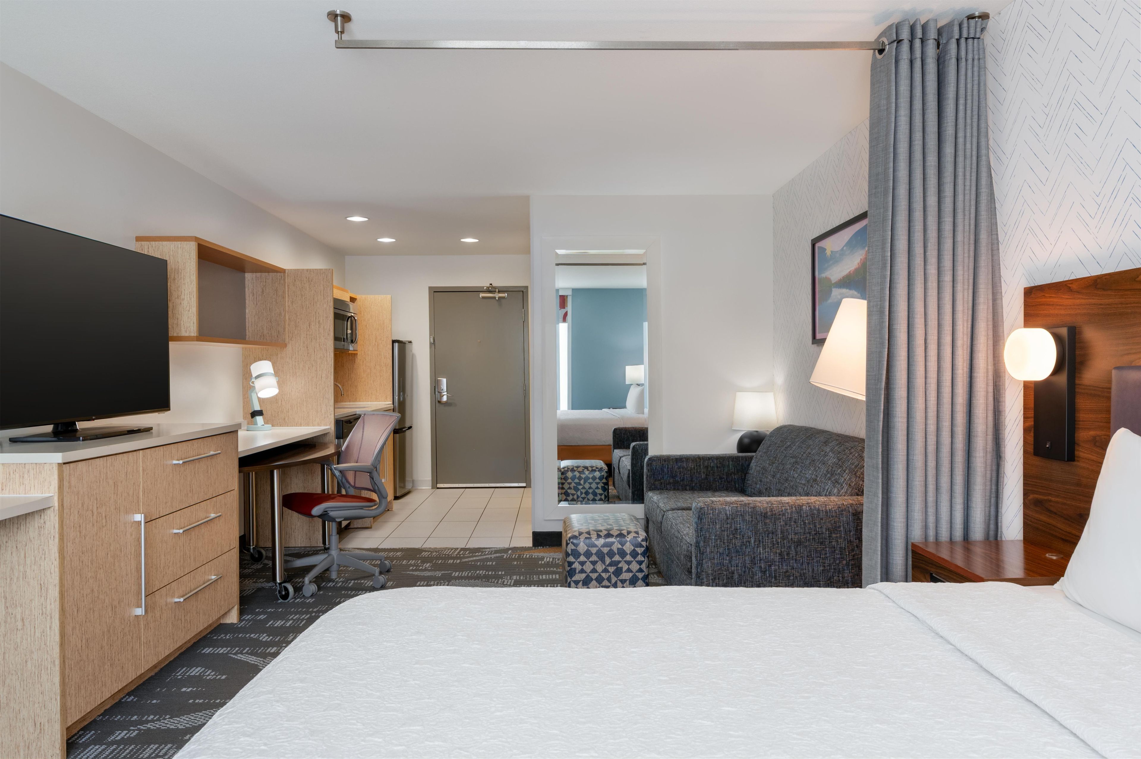 Habitación de hotel con cama, escritorio con computadora, televisor, sofá y baño visible a través de una puerta abierta.